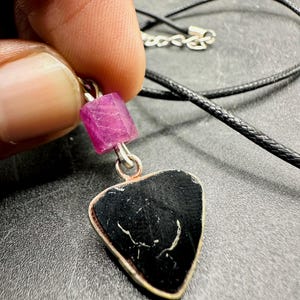 Op de afbeelding: Een handgemaakte ketting met een zwarte, hartvormige hanger en een zilverkleurige rand. Een kleine, roze, rechthoekige edelsteen is met een zilverkleurige lus aan de hanger bevestigd. De ketting heeft een zwart koord.
