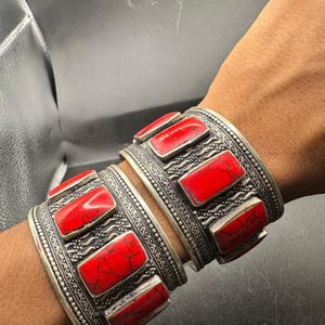 May include: Two silver-toned cuff bracelets with rectangular red stone inlays. The bracelets have intricate etched patterns and are worn on a wrist. The jewelry is likely handmade and has a vintage or ethnic aesthetic.