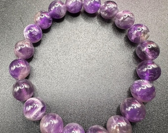 May include: A bracelet of polished amethyst beads, each a deep purple with translucent patches. The beads are strung together, forming a circular shape. The bracelet is set against a dark background.