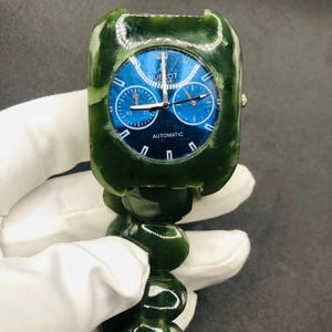 Op de afbeelding: Een groene jade horloge met een blauwe wijzerplaat en het woord "AUTOMATIC" onderaan. Het horloge heeft een vierkante wijzerplaat en een jade band gemaakt van grote, ronde kralen. Het horloge wordt vastgehouden door een gehandschoende hand.