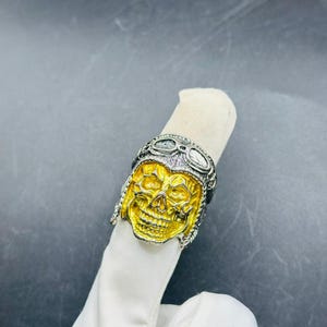 Könnte beinhalten: Ein silberner und goldener Totenkopfring mit detailliertem Design. Der Totenkopf hat eine goldene Oberfläche, und der Ring hat ein silbernes Fliegerhelm-Design. Der Ring wird auf einer weißen Oberfläche gezeigt.