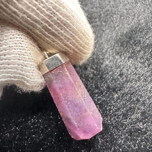 Op de afbeelding: Een gefacetteerde, zeshoekige roze edelsteen hanger met een zilverkleurige metalen kap. Het kristal vertoont natuurlijke insluitingen en kleurvariaties, variërend van diep magenta tot lichtere lavendeltinten, hangend aan een metalen oogje.