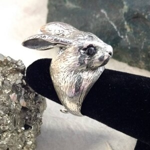 May include: A silver ring featuring a detailed rabbit head design with black eyes. The ring is displayed on a black velvet ring holder.