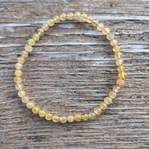 May include: A beaded bracelet composed of small, round, translucent yellow-orange gemstones. The bracelet is strung on a thin, elastic cord and rests on a wooden surface. The gemstones have a polished appearance, reflecting light.