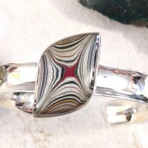 May include: A silver cuff bracelet with a colorful agate gemstone set in the center. The gemstone has a swirling pattern of black, white, red, and gold.