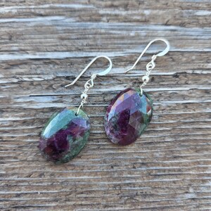 May include: A pair of gemstone earrings featuring faceted, oval-shaped ruby stones. The stones show a mix of deep red and green hues, suspended from silver ear wires. The earrings are displayed on a wooden surface.