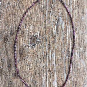 May include: A delicate necklace composed of small, round, dark pink beads. The beads are strung together, forming a single strand that fastens with a silver clasp. The necklace rests on a rustic, wooden surface, showcasing its natural beauty.