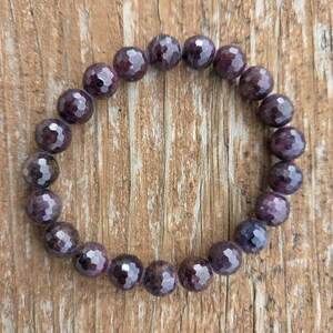 May include: A faceted, dark purple gemstone bracelet. The beads are round and have a multi-faceted surface, creating a sparkling effect. The bracelet is strung on an elastic band, and is displayed on a wooden surface.