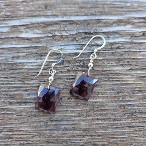 May include: Silver drop earrings with faceted, dark purple gemstones. The earrings have a simple design with a small silver bead and a hook closure. The gemstones are rectangular and have a polished finish.