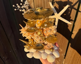 May include: A decorative seashell arrangement featuring a variety of shells, including conch and scallop shells, accented with gold paint and faux pearls. A white starfish and small white shells are also included. The arrangement is held in a hand.