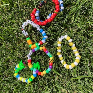 May include: Four colourful beaded bracelets with letters and various colours, including red, blue, yellow, and green, are displayed on green grass. Some bracelets have letters spelling out names. The bracelets are a fun and playful accessory.