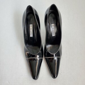 May include: A pair of black patent leather court shoes with a pointed toe and a distinct white stitched seam across the vamp. The interior features a white label with the text "the saddler" and "MADE IN ITALY" printed in black on the black insole.