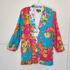 May include: A long-sleeve blazer featuring a vibrant floral pattern in shades of hot pink, bright turquoise, yellow, and orange. The garment has a white notched lapel, two white buttons for closure, and two front pockets. A Karen Kane label is visible.