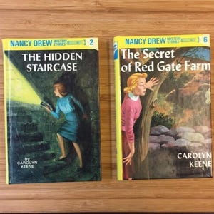 Peut inclure: Deux livres Nancy Drew Mystery Stories intitulés The Hidden Staircase et The Secret of Red Gate Farm par Carolyn Keene. La première couverture montre une personne avec une lampe torche dans un escalier, la seconde près d'un arbre.