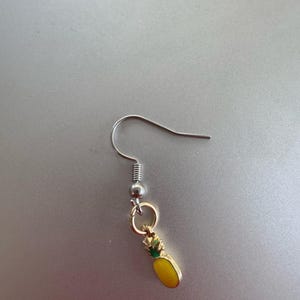 May include: A single silver-tone earring with a small, gold-colored pineapple charm. The pineapple has a yellow body and green top. The earring hook is a simple, curved design.