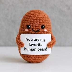 Puede incluir: Una figura de frijol marrón tejida a ganchillo con una cara sonriente, sosteniendo un letrero blanco que dice "You are my favorite human bean!". La figura tiene brazos pequeños y mejillas rosadas. El fondo es gris suave.