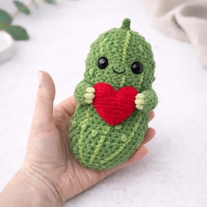 Puede incluir: Una muñeca de pepinillo de ganchillo verde hecha a mano con una cara sonriente y un corazón rojo. El pepinillo tiene ojos negros y brazos pequeños. Un artículo hecho a mano encantador y caprichoso.