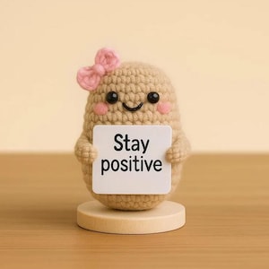 Puede incluir: Una figura de crochet con forma de patata con un lazo rosa y mejillas rosadas, sosteniendo un cartel que dice "Stay positive". La figura está sobre una pequeña base de madera, sobre un fondo neutro. El diseño general es lindo y caprichoso.