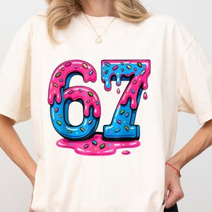 Könnte beinhalten: Cremefarbenes T-Shirt mit einer farbenfrohen Grafik der Zahl 67. Die Zahlen sind blau mit rosa Zuckerguss und Streuseln, die einem Donut ähneln. Der Zuckerguss tropft herunter und erzeugt ein verspieltes und süßes Design.