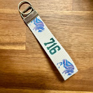 Op de afbeelding: Een witte stoffen sleutelhanger met het nummer "716" in groene en blauwe patronen. Het heeft een zilverkleurige metalen sluiting en ring. De achtergrond is een houten oppervlak.