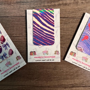May include: Three magnetic bookmarks with unique designs. One features footballs and bows, another has a zebra stripe pattern, and the third showcases footballs and red shapes. Each bookmark is on a card with text: "MAGNETIC BOOKMARK CUSTOM CRAFT LAB BY DEE."
