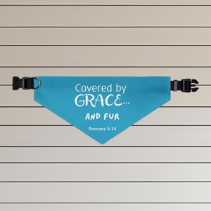 May include: A turquoise dog bandana with the text "Covered by GRACE... AND FUR" and the Bible verse Romans 6:14 in white. The bandana has a black adjustable buckle and a D-ring.