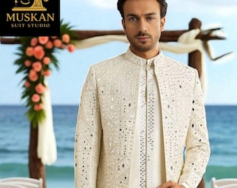 May include: A cream-coloured suit with detailed mirror embellishments. The jacket has a mandarin collar and long sleeves. The suit is worn over a matching tunic and trousers. The background shows a beach wedding setting with floral decorations and the text "MUSKAN SUIT STUDIO".