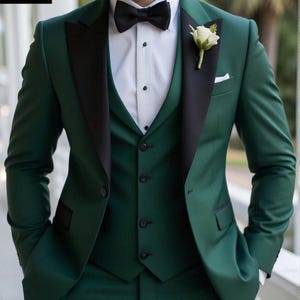 May include: A three-piece emerald green suit with a black lapel and a black bow tie. The suit includes a jacket, vest, and trousers. A white flower is pinned to the lapel. The suit is from Muskan Suit Studio.