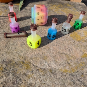 Könnte beinhalten: Eine Sammlung farbenfroher Würfel und flaschenförmiger Behälter. Die Würfel umfassen einen regenbogenfarbenen D20 und mehrere kleine, nummerierte Flaschen in verschiedenen Farben wie Blau, Grün und Gelb. Ein brauner, zylindrischer Gegenstand ist ebenfalls vorhanden.