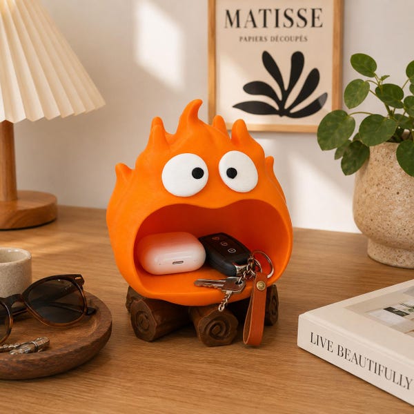 May include: A bright orange, flame-shaped storage organizer with large white eyes and a wide open mouth. The figure sits on a base designed to look like brown wooden logs. Inside the mouth, a white earbud case and a set of keys are stored.