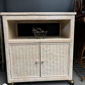 May include: Off-white woven wicker cabinet with two doors, an open shelf, and a top shelf. The cabinet has a rectangular shape and sits on small brass casters. A decorative metal basket sits on the open shelf.
