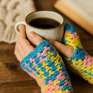 Puede incluir: Un par de guantes sin dedos de ganchillo coloridos en azul, amarillo y rosa. Los guantes se usan mientras se sostiene una taza de cerámica moteada llena de líquido oscuro. El fondo incluye una superficie de madera y un libro.
