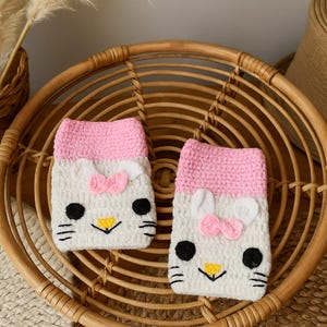 Puede incluir: Dos fundas de teléfono de ganchillo hechas a mano con un diseño de Hello Kitty. Las fundas son blancas con detalles en rosa, incluido un lazo rosa, y rasgos faciales negros y amarillos. Se exhiben en una cesta de mimbre.