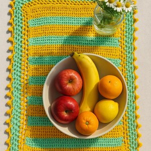 May include: A rectangular, handmade crochet placemat with yellow and green stripes and a scalloped edge. A bowl of fruit, including red apples, a banana, oranges, and a lemon, sits on the placemat. A small vase of white daisies is also present.
