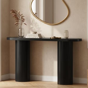May include: A black console table with a rounded top and two cylindrical, fluted legs. The table is adorned with decorative items, including a vase with dried plants, books, and a gold-framed mirror. The room has a neutral color palette.