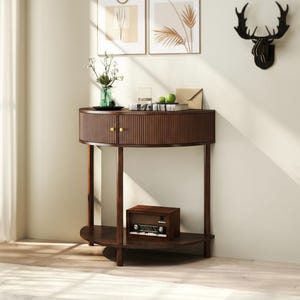 May include: A dark brown, half-moon console table with a ribbed front and two gold-toned knobs. The table has a lower shelf and is supported by four slender legs. A vintage radio sits on the lower shelf.