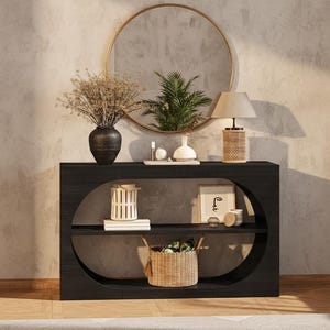 May include: A black console table with oval cutouts, holding decorative items. A gold-framed mirror hangs above. Items include a vase with dried flowers, a lamp, a white vase, a framed print, candles, and a woven basket.