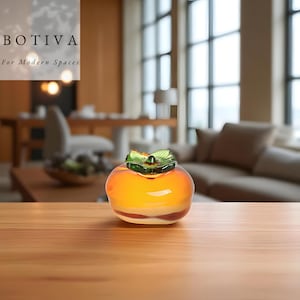 May include: A decorative glass persimmon with a green leaf design on top. The fruit is a mix of orange, yellow, and white, and sits on a wooden surface. The image includes the text "BOTIVA For Modern Spaces".