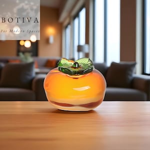 May include: A glass persimmon fruit sculpture with an orange body and green leaf detail. The sculpture is displayed on a wooden surface. The text "BOTIVA For Modern Spaces" is visible in the background.