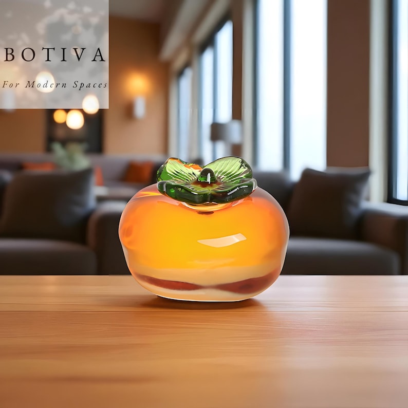 May include: A glass persimmon fruit sculpture with an orange body and green leaf detail. The sculpture is displayed on a wooden surface. The text "BOTIVA For Modern Spaces" is visible in the background.