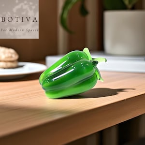 Puede incluir: Una figura de pimiento verde de cristal brillante. La superficie lisa refleja la luz, resaltando sus suaves curvas y detalles realistas. La figura está colocada sobre una superficie de madera. El logotipo "BOTIVA For Modern Spaces" está en la esquina superior izquierda.