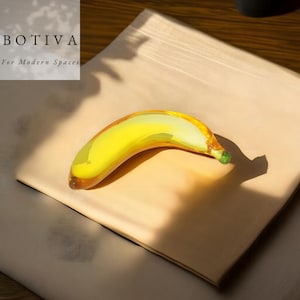 May include: A glass banana sculpture in shades of yellow, green, and brown sits on a beige surface. The banana is curved and detailed, with a green tip. The image includes the text "BOTIVA For Modern Spaces".