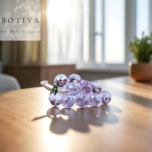 Puede incluir: Un racimo de uvas decorativo de cristal con orbes facetados de color púrpura claro. Las uvas están unidas a un tallo transparente con hojas verdes. La imagen incluye el texto "BOTIVA For Modern Spaces".