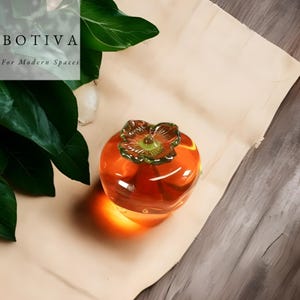 May include: A clear, orange glass persimmon fruit sculpture with a green and yellow flower-like top. The sculpture is on a light brown surface. The text "BOTIVA For Modern Spaces" is in the upper left corner.
