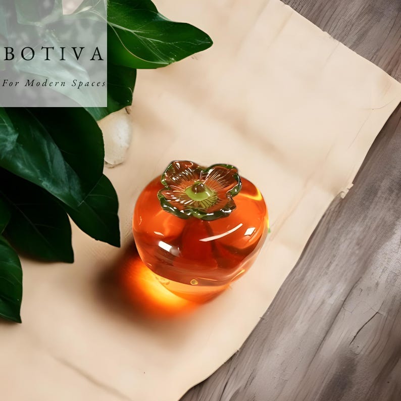May include: A clear, orange glass persimmon fruit sculpture with a green and yellow flower-like top. The sculpture is on a light brown surface. The text "BOTIVA For Modern Spaces" is in the upper left corner.