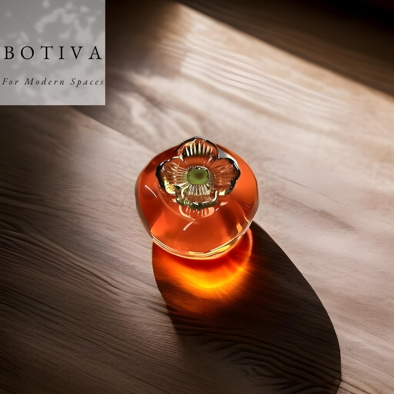 May include: A decorative glass object with a flower-shaped top and a round, orange-coloured body. The object is on a wooden surface. The text "BOTIVA For Modern Spaces" is in the upper left corner.
