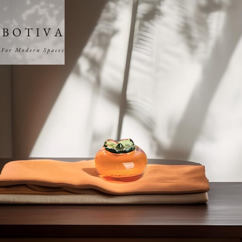 May include: A decorative glass persimmon in orange, with a green leaf-like top. It rests on folded orange and beige cloths, on a dark brown surface. The text "BOTIVA For Modern Spaces" is in the upper left corner. The persimmon is a decorative item.