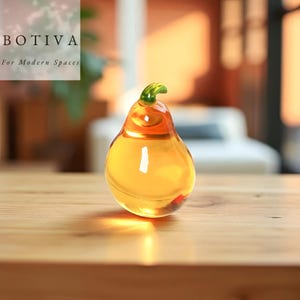 Puede incluir: Una pera decorativa de cristal con un tallo verde y un cuerpo amarillo-naranja. La pera está sobre una superficie de madera. El texto "BOTIVA For Modern Spaces" está en la esquina superior izquierda.