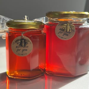 Puede incluir: Dos frascos de vidrio llenos de miel dorada. Cada frasco tiene una tapa dorada y una etiqueta redonda con un diseño de abeja y las palabras "with love for you". La miel es de color ámbar intenso.