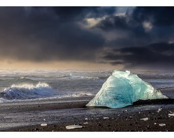 Puede incluir: Fotografía de paisaje de una playa de arena negra con un gran iceberg azul translúcido en la orilla. Las olas del océano rompen contra la playa bajo un cielo dramático y nublado.